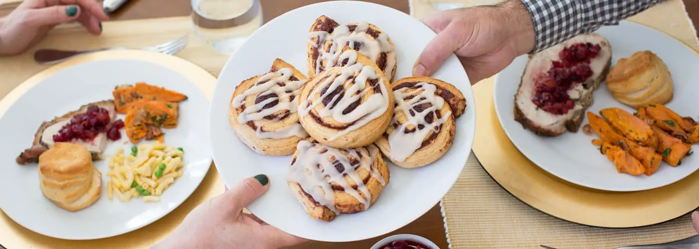 Hands passing a plate of cinnamon rolls across a dinner table.