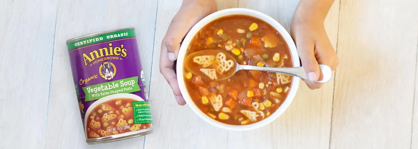 A child's hands wrapped around a bowl of soup next to a can of Annie's Organic Vegetable Soup With Farm Shaped Pasta.
