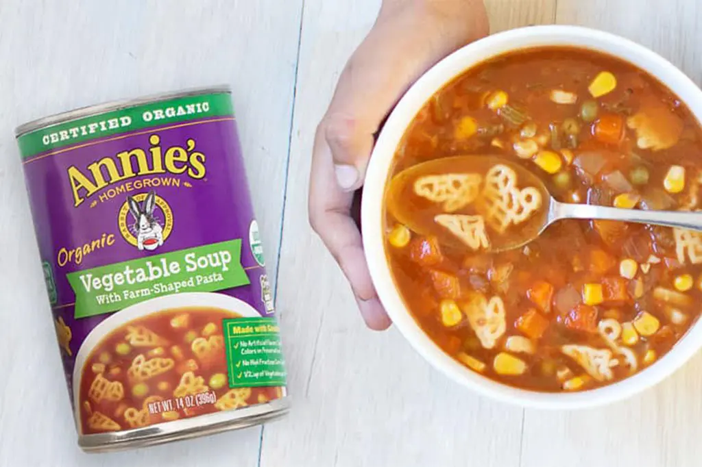 A child's hands wrapped around a bowl of soup next to a can of Annie's Organic Vegetable Soup With Farm Shaped Pasta.