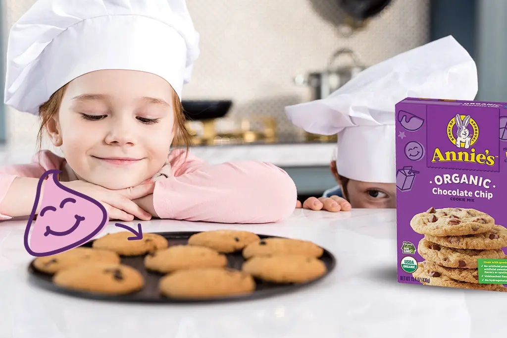 Children in chef's hats lean over a kitchen counter eyeing up their freshly baked chocolate chip cookies next to a box of Annie's Organic Chocolate Chip Cookie Mix.