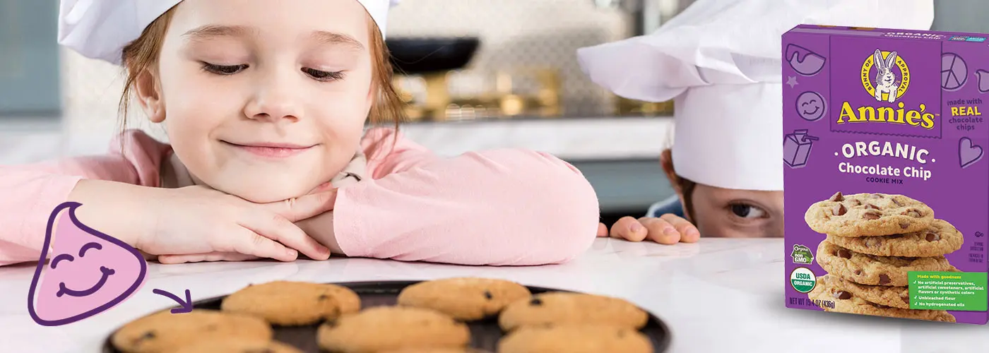 Children in chefs' hats peering over the counter at their freshly baked chocolate chip cookies next to a box of Annie's Organic Chocolate Chip Cookie Mix.