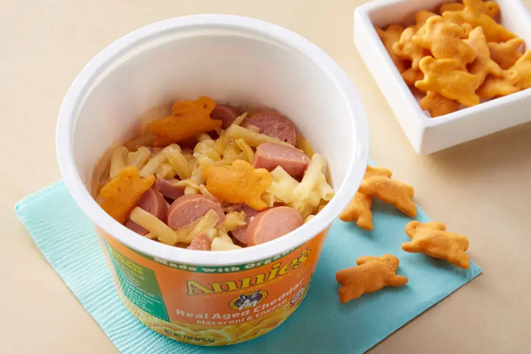 Cheesy Hot Dog Macaroni & Cheese recipe in a single-serving cup of Annie's Real Aged Cheddar Microwavable Macaroni & Cheese cup sitting on a teal napkin on a surface with single Cheddar Bunnies laying around it.