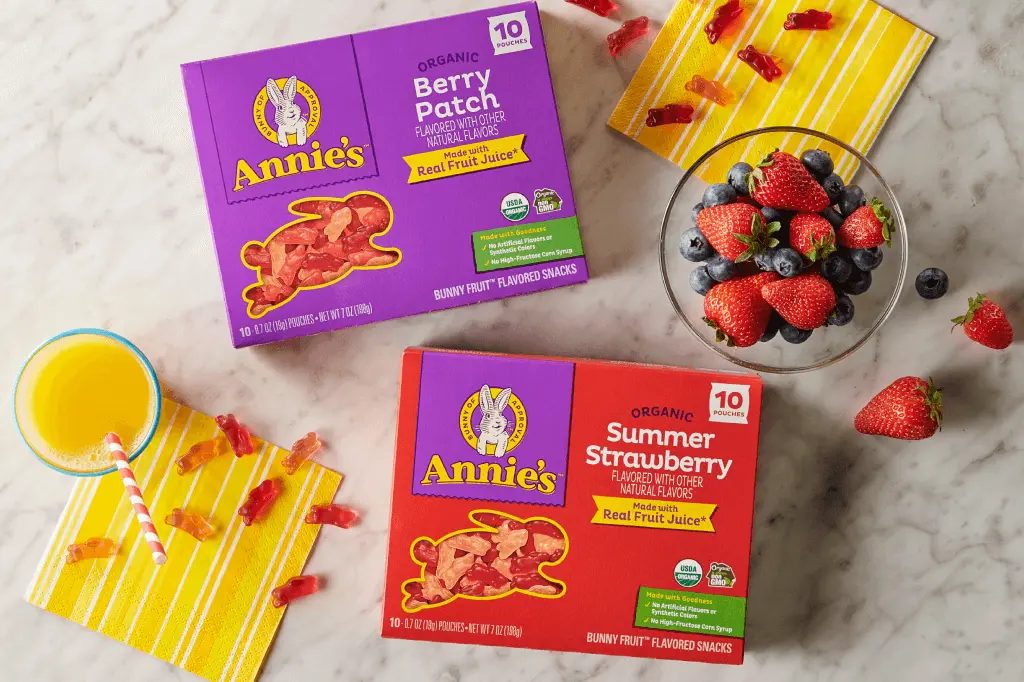 Single boxes of Annie's Organic Summer Strawberry and Annie's Organic Berry Patch Fruit Snacks on a marble table with a bowl of fruit and orange juice.
