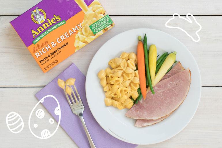A plate of Annie's Shells and Aged Cheddar, ham, and a variety of vegetables next to a box of the same and a fork with three shells, all on an Easter themed background