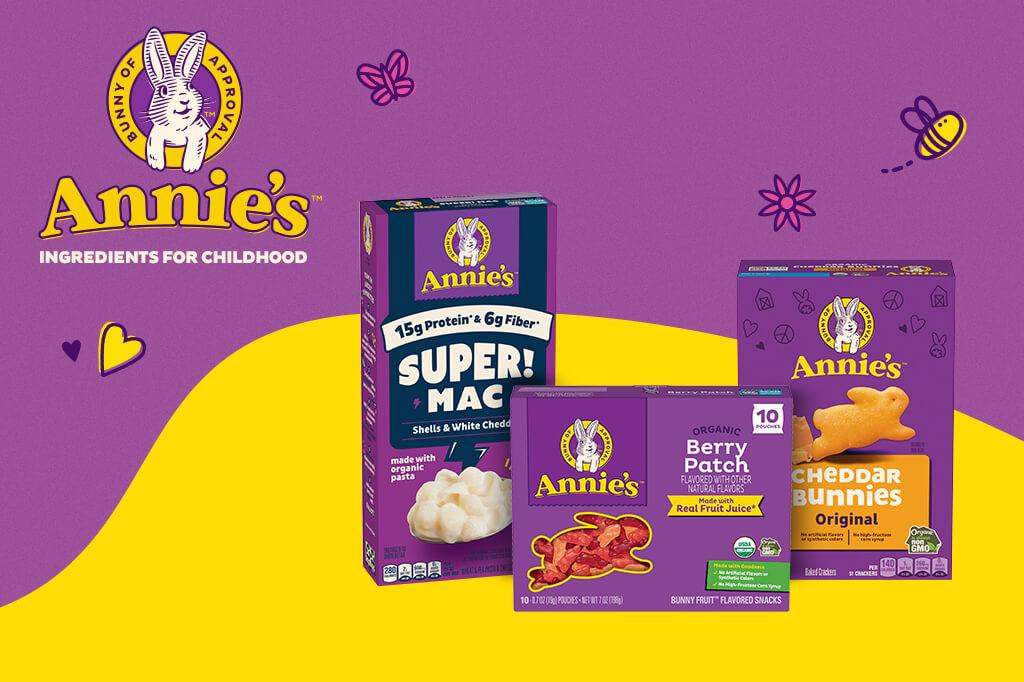 Annie's brand promotional image featuring three product boxes: Super Mac Shells & White Cheddar, Organic Berry Patch Bunny Fruit-Flavored Snacks, and Cheddar Bunnies Original Baked Crackers. The Annie's logo with a rabbit and the text 'Bunny of Approval' is visible, along with the slogan 'Ingredients for Childhood.' Decorative elements include a bee, heart, and butterfly illustrations.