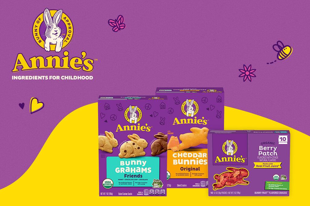 Annie's product lineup featuring three boxes. From left to right: Bunny Grahams Friends, Cheddar Bunnies Original, and Organic Berry Patch fruit-flavored snacks. The boxes display the 'Bunny of Approval' logo and highlight features like USDA Organic and real fruit juice. The background includes playful illustrations of a heart, butterfly, and bee.