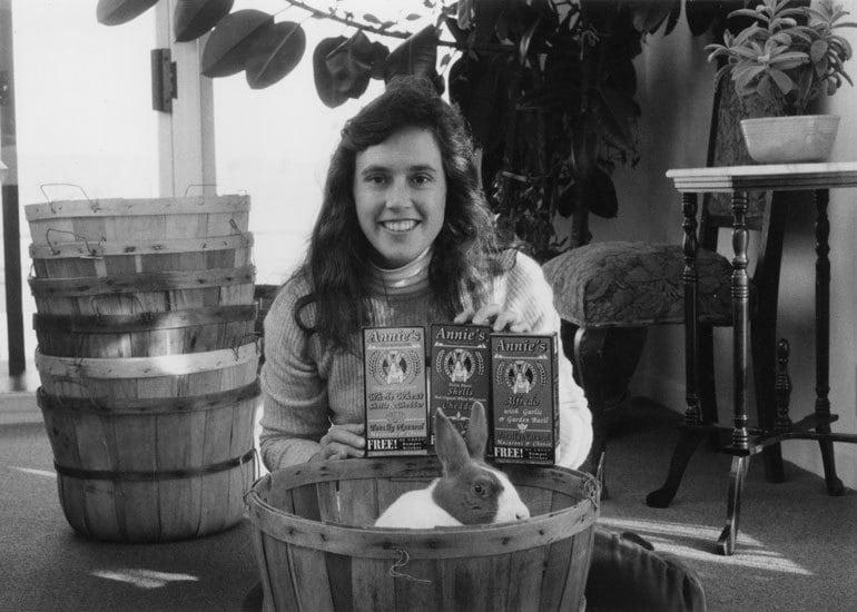 Black and white image of Annie Withey holding three boxes of Annie's Mac and Cheese. Her pet rabbit sits in a basket in front of her.