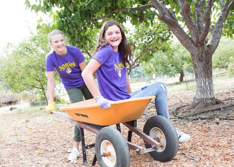 Two pre-teen girls laughing, while one pushes the other in a wheelbarrow.