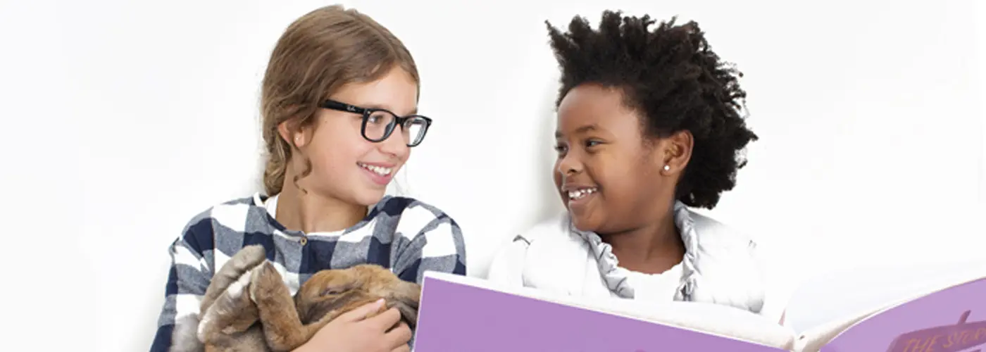 Two children smiling at each other, one holding a real bunny and the other holding a book.