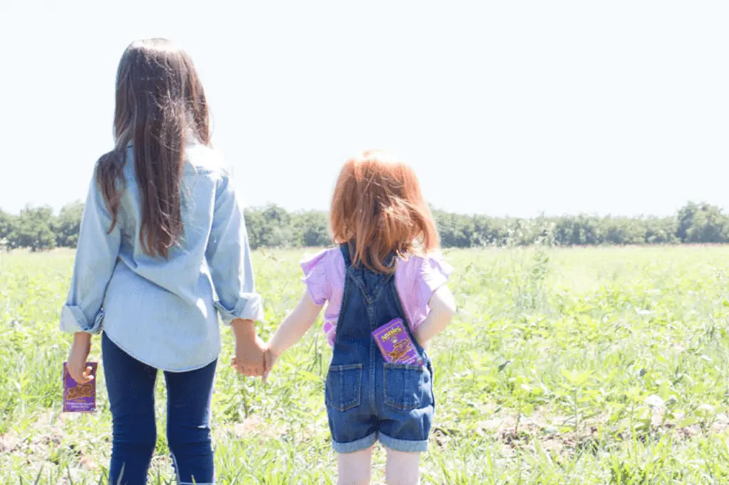 Two young girls in a field, holding hands. One girl is holding a individual sized fruit snacks and the other has one in her back pocket.