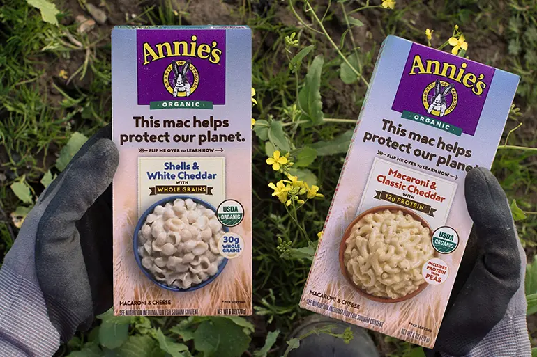 Hands wearing gardening gloves holding two boxes of Annie's macaroni and cheese.
