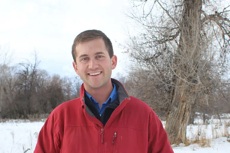 A smiling man standing in a snowy field with bare trees in the background.