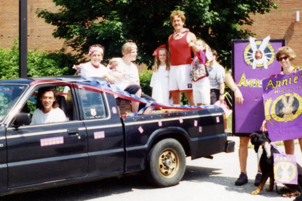 An old photograph of people piled into a car with others wearing cardboard slats with Annie's branding.