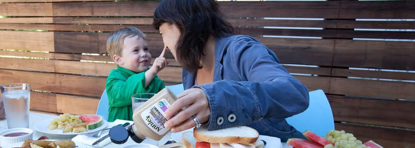 An outdoor lifestyle image of a mother holding a bottle of Annie's Organic Dijon Mustard at a dinner table while a little boy touches her nose.