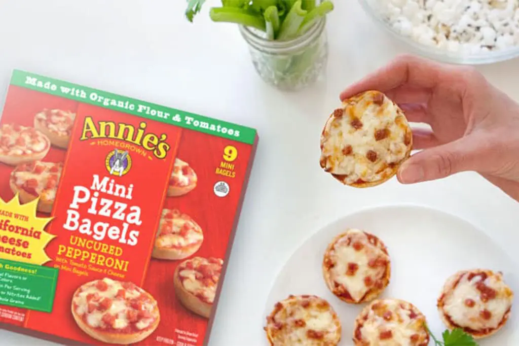 A box of Annie's Mini Pizza Bagels Uncured Pepperoni sits on a table next to a hand picking up a single mini pizza bagel from a plate of five mini pizza bagels.