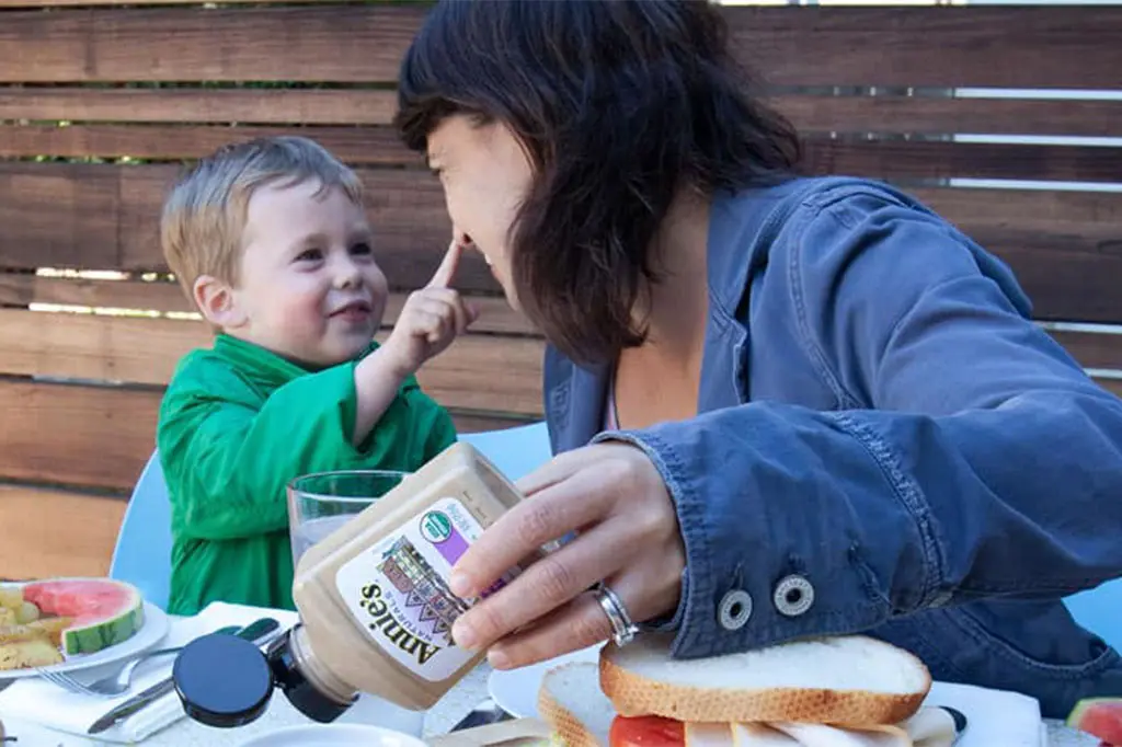 An outdoor lifestyle image of a mother holding a bottle of Annie's Organic Dijon Mustard at a dinner table while a little boy touches her nose.