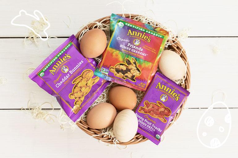 A basket containing five eggs and three packages of different Annie's snacks on an Easter themed background