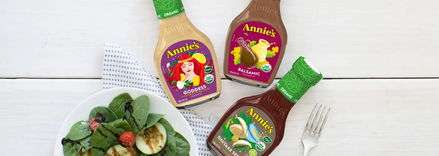 A single bottle of Annie's Organic Goddess Dressing, Annie's Organic Balsamic Vinaigrette and Annie's Organic Shiitake Sesame Vinaigrette next to a plate of spinach salad and a fork.