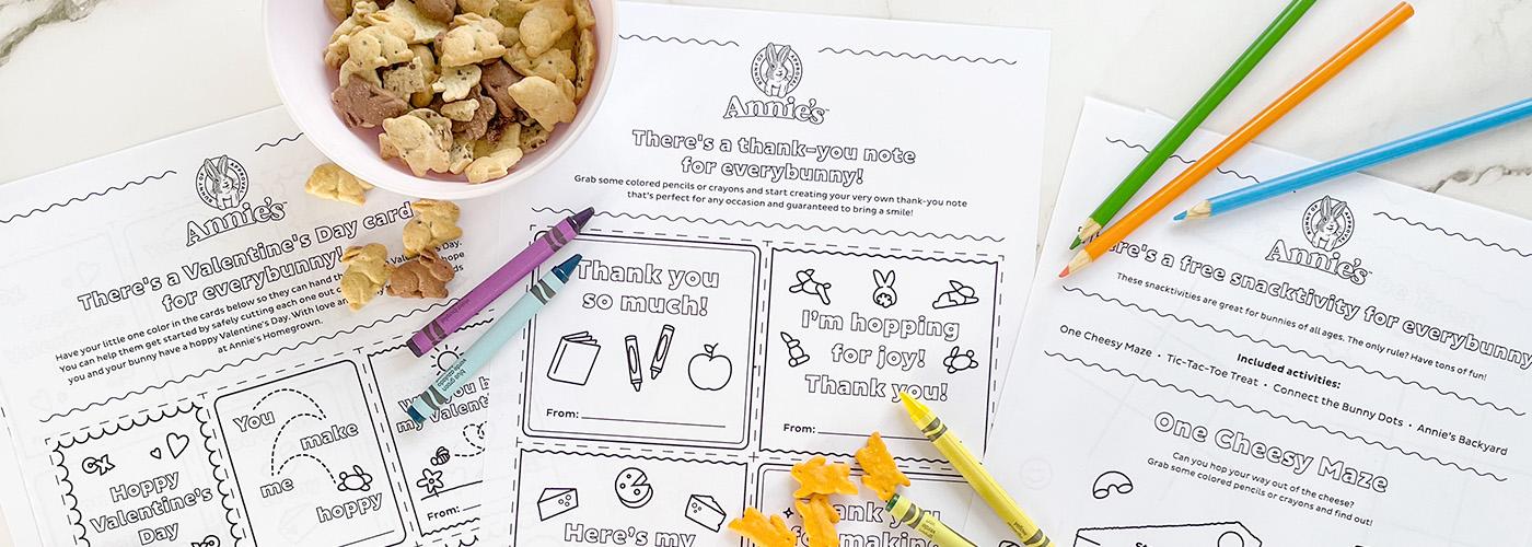 Three different Annie's activity sheets laying out with single Cheddar Bunnies sprinkled around, Bunny Grahams in a bowl, and a few colored pencils and crayons.