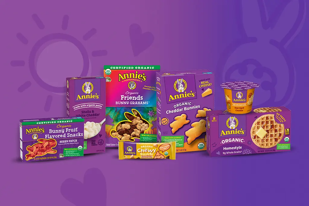 A grouping of Annie's packaging, including a box of fruit snacks, mac and cheese, Bunny Grahams, Cheddar Bunnies, waffles and single serving of a granola bar and microwave mac and cheese on a purple background.