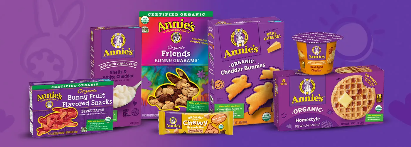 A grouping of Annie's packaging, including a box of fruit snacks, mac and cheese, Bunny Grahams, Cheddar Bunnies, waffles and single serving of a granola bar and microwave mac and cheese on a purple background.