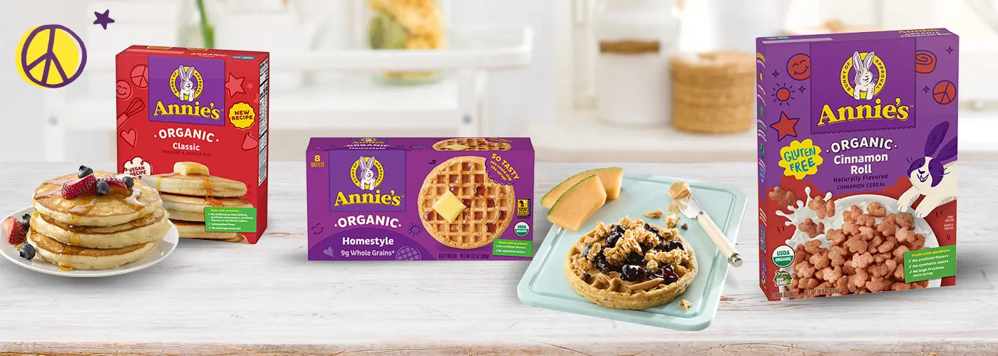 A plate of pancakes, a box of Annie's Organic Classic Pancake, a box of Annie's Organic Homestyle Waffles next to a plate with a waffle and a box of Annie's Organic Cinnamon Roll Cereal on a kitchen counter.