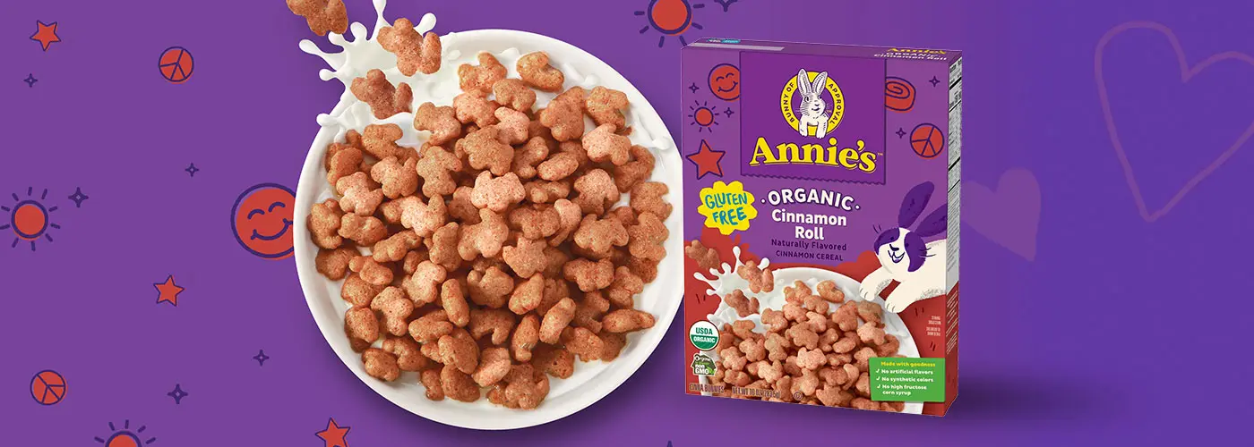 A bowl of Annie's Organic Cinnamon Roll Cereal next to a white bowl of Cinnamon Roll cereal and milk on a purple background.