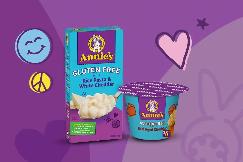Single box of Annie's Gluten Free Rice Pasta And White Cheddar next to a single cup of Annie's Gluten Free Real Aged Cheddar single serving cup.