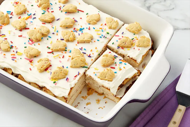 A purple pan of Birthday Cake Bunnies Cinnamon Roll Lasagna with frosting, sprinkles and Annie's Organic Birthday Cake Bunny Grahams Baked Graham Snacks.