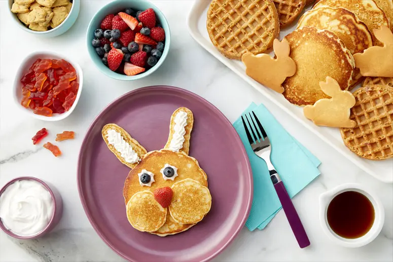 A bunny face made with pancakes, whipped cram, blueberries for eyes and a strawberry nose on plate surrounded by other toppings and a platter with Annie's waffles, regular pancakes and small bunny shaped pancakes.