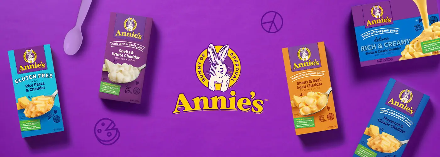 Purple background with the Annie's logo in the middle with five front of pack products of mac and cheese products with a spoon.