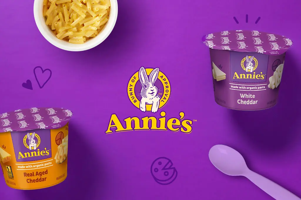 Purple background with the Annie's logo in the middle and two front of pack products of mac and cheese microwave cup products with a spoon.