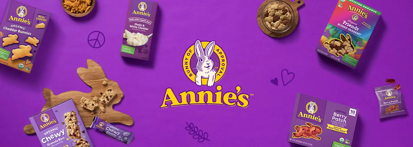Purple background with Annie's logo in the middle and some front of pack snack products and bowls of Annie's Cheddar Bunnies and Bunny Grahams.