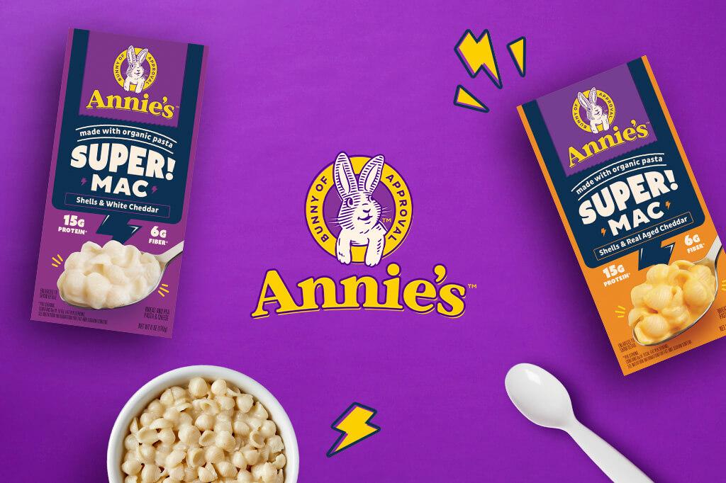 Annie's logo on a purple background with packages of Super Mac and a bowl of Shells And White Cheddar and a spoon.