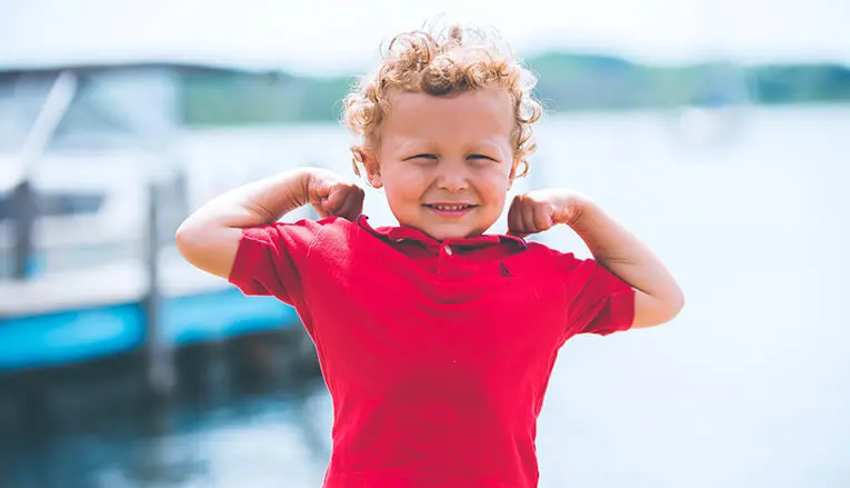 A little boy in a red shirt flexing his muscles with a lake in the background.
