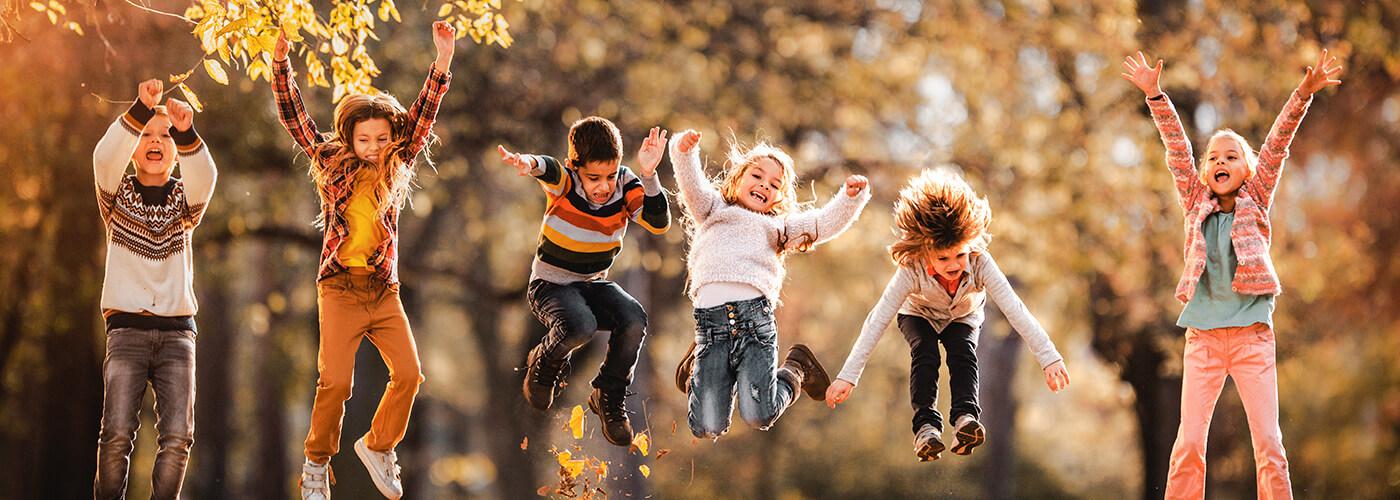A group of six kids jumping in the air with fall leaves in the foreground and background.
