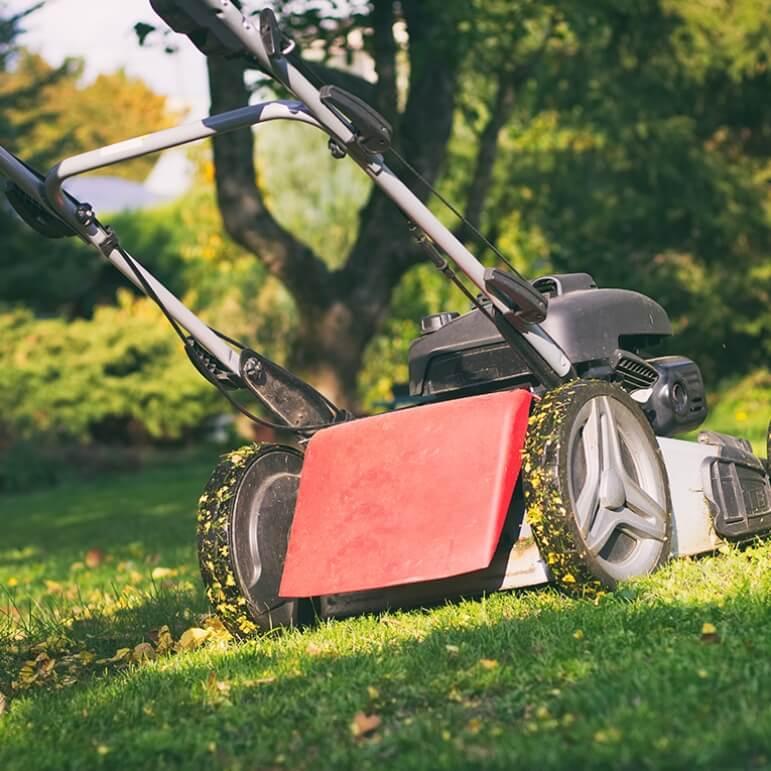 A close up of a red lawn mower on a green grass lawn with grass around it's wheels.