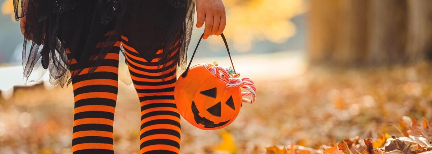 A close-up of the back of someone's legs wearing orange and black strip leggings, a black skirt, and holding an orange Halloween bucket filled with candy walking outside with leaves the ground.