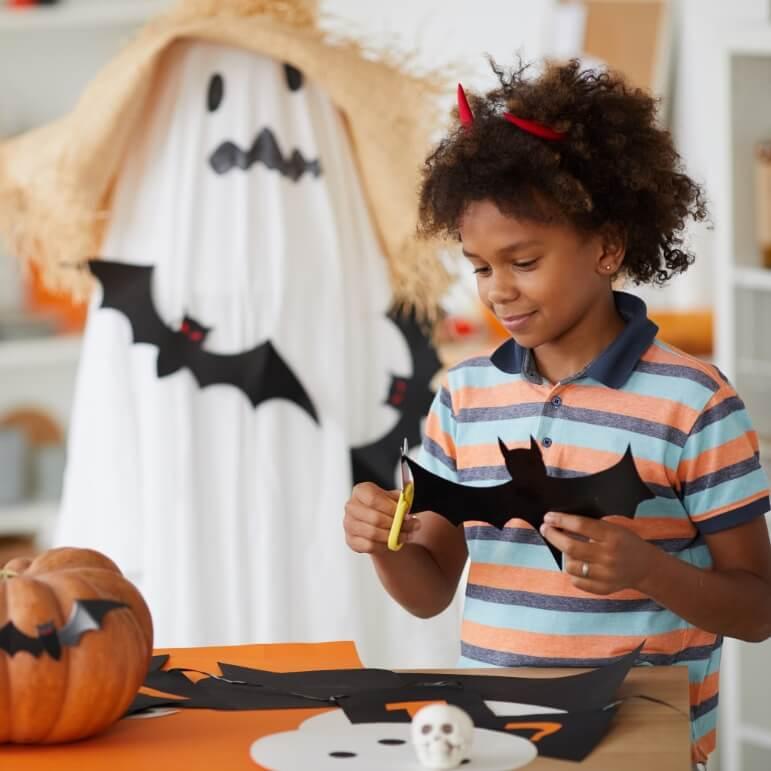 A child cutting out black bats with scissors and a white ghost decoration in the background.