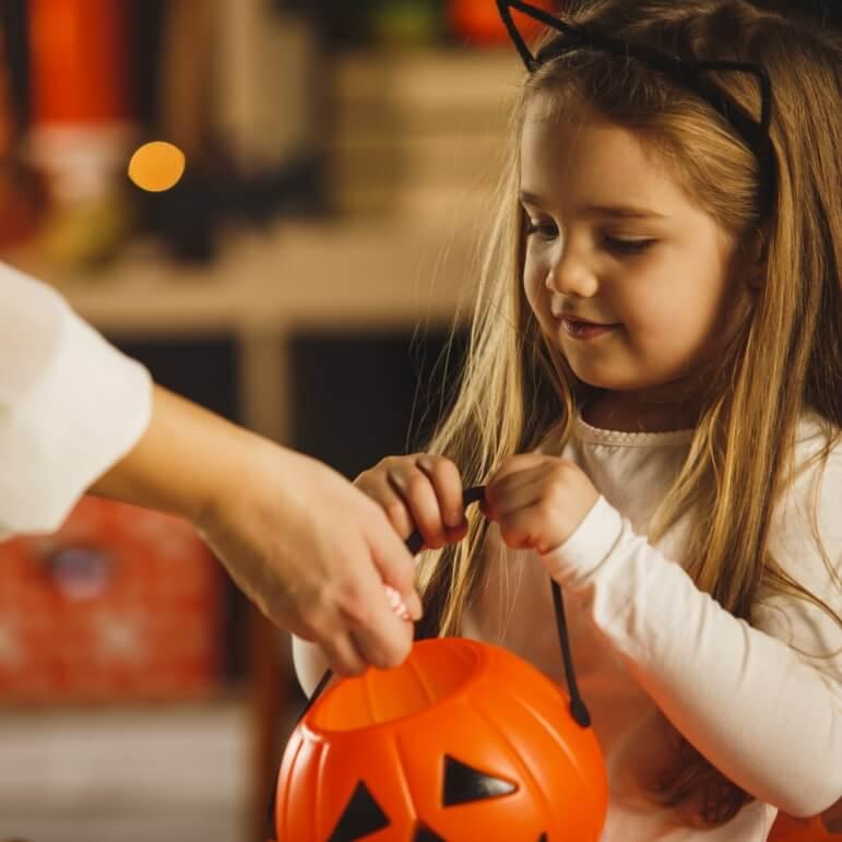 A little girl with cat ears on holding a pumpkin bucket with a hand putting a treat into it.