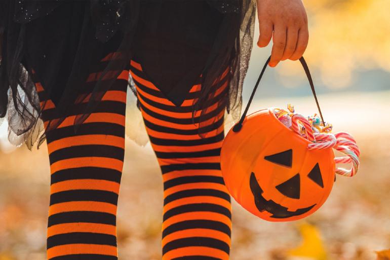 A close-up of the back of someone's legs wearing orange and black strip leggings, a black skirt, and holding an orange Halloween bucket filled with candy walking outside with leaves the ground