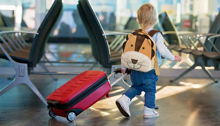 Little child in a airport with a dog backpack on and rolling a red suitcase.
