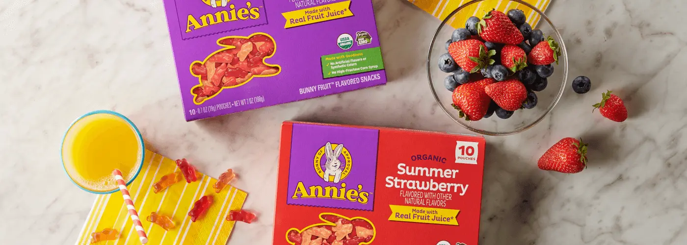 Single boxes of Annie's Organic Summer Strawberry and Annie's Organic Berry Patch Fruit Snacks on a marble table with a bowl of fruit and orange juice.
