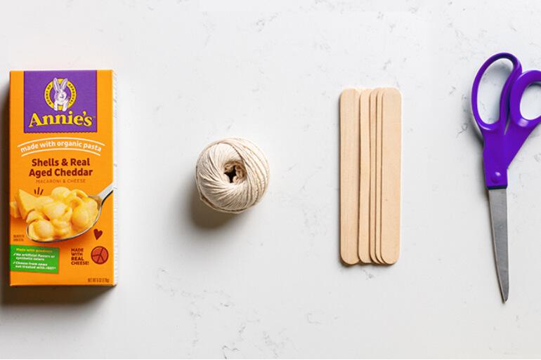 A box of Annie's Shells & Real Aged Cheddar, a ball of string, craft sticks and a pair of purple scissors arranged in a line