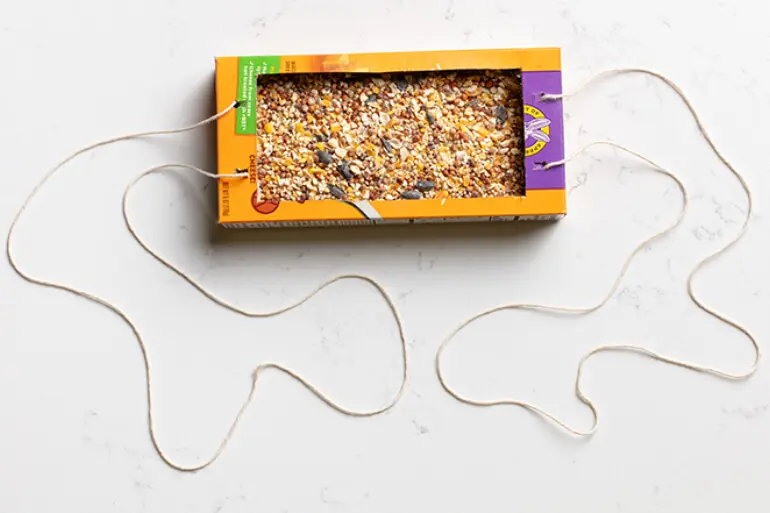 A Annie's box laying on it's side with the top cut open filled with bird seed and string-tied to either end.