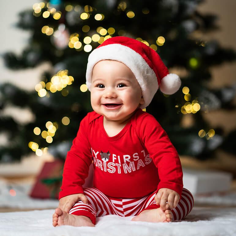A baby wearing a red shirt and a Santa hat is seated on the floor with a Christmas tree behind them