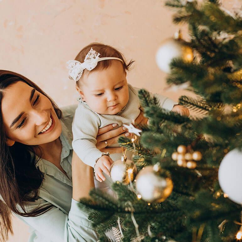 A woman smiling holding a baby decorating a Christmas tree