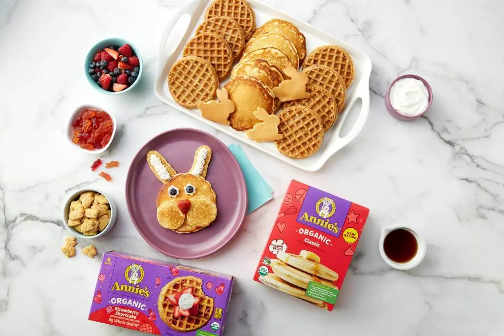 Assorted breakfast foods including a bunny made out of waffles and pancakes. Includes a a cup of fruit, bunny grahams, and fruit snacks. Also includes the top portion of a front of box of an Annies Pancakes and Waffles in the corner