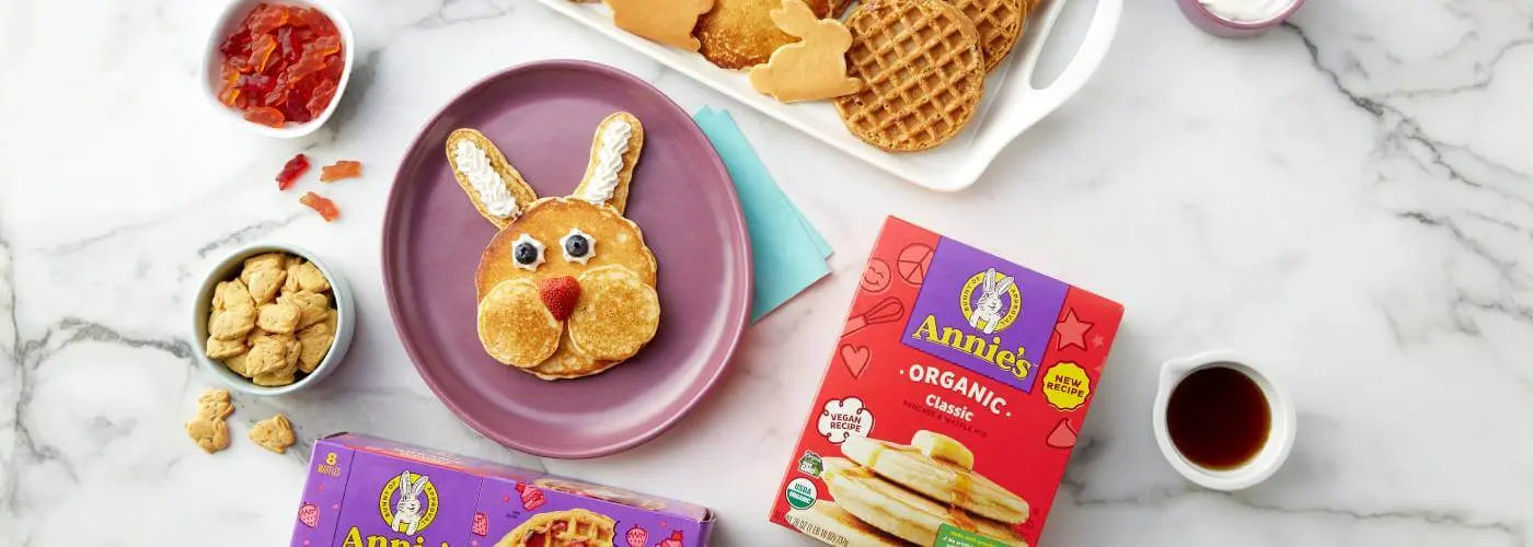 Assorted breakfast foods including a bunny made out of waffles and pancakes. Includes a a cup of fruit, bunny grahams, and fruit snacks. Also includes the top portion of a front of box of an Annies Pancakes and Waffles in the corner