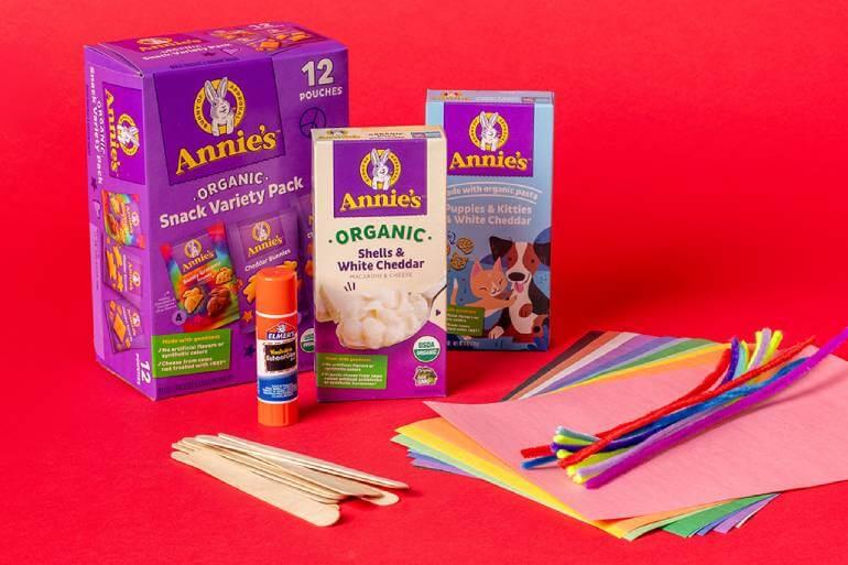 Craft supplies: paper, scissors, a glue stick, wooden glue sticks, multicolored pipe cleaners, arranged in front of a Annies Snack Variety Pack, Organic Shells & White Cheddar Mac & Cheese and Puppies & Kitties White Cheddar Mac & Cheese boxes.